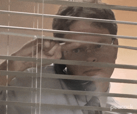 A man wearing a shirt and tie is seen looking through window blinds in a gif from the popular TV show The Office.